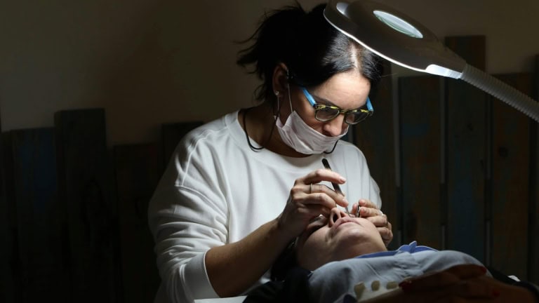 A woman wearing a face mask and glasses performs an eye lash service on a woman lying down