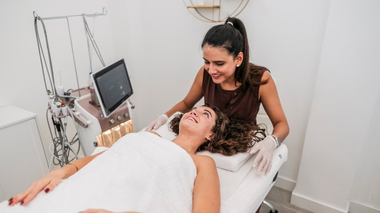 A skincare professional wearing gloves smiles down at a client lying on a treatment bed next to a piece of medical equipment.