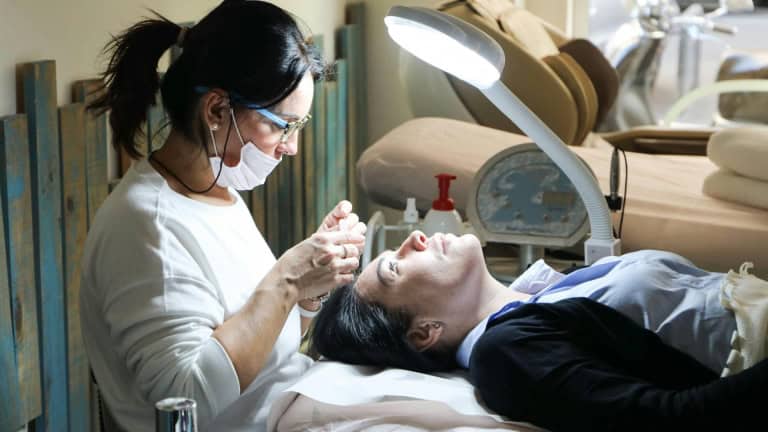 A person wearing a mask and glasses works on another person's eyelashes under a bright lamp.